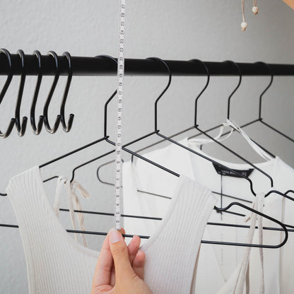 Eine Person hält ein Maßband senkrecht an Kleidung auf Kleiderbügeln in einem Kleiderschrank. Auf den Kleiderbügeln hängen helle Kleidungsstücke, und auf der linken Seite sind mehrere leere schwarze Kleiderbügel-Bündel aus stabilem Metall von Rod & Knot zu sehen. Der Hintergrund des Kleiderschranks ist eine neutrale graue Wand.